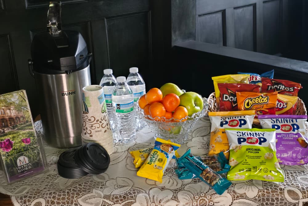 A table displays a coffee maker, bottled water, a bowl of fruit, and various snacks including chips and popcorn.