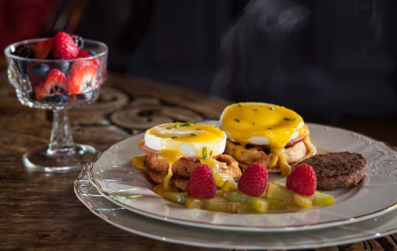 A plate of eggs Benedict with two poached eggs and hollandaise sauce, accompanied by sausage and a side of mixed berries in a glass dish.