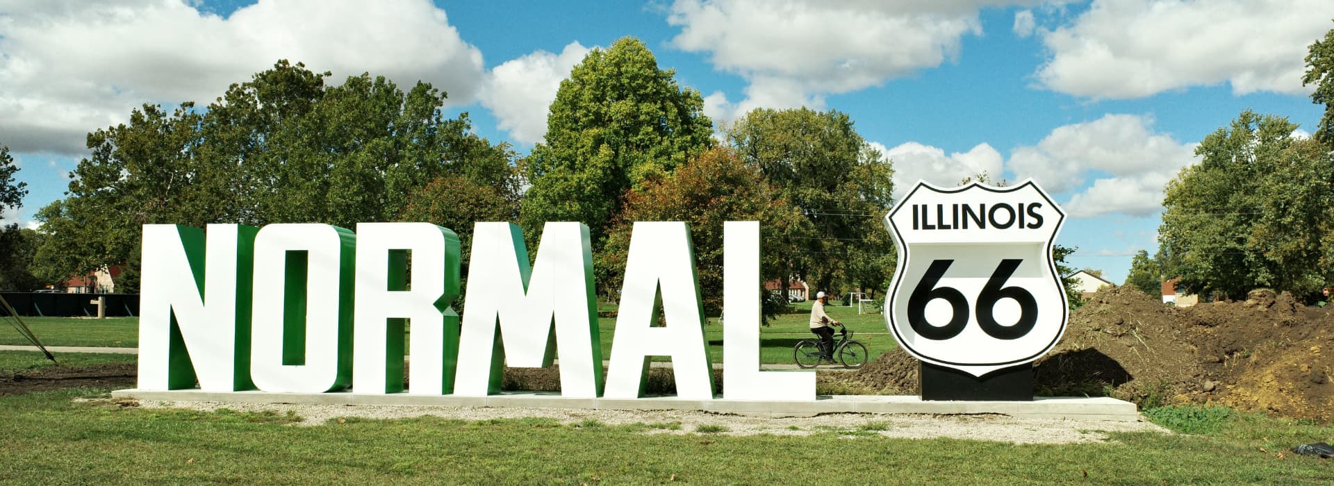 A large "NORMAL" sign alongside a Route 66 marker in a park with trees and a cyclist in the background.