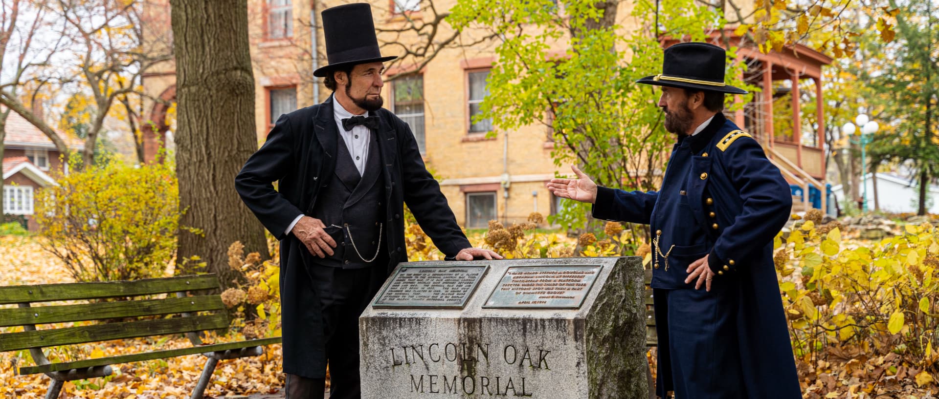 Two actors in period costumes stand by a memorial plaque in a park during autumn.