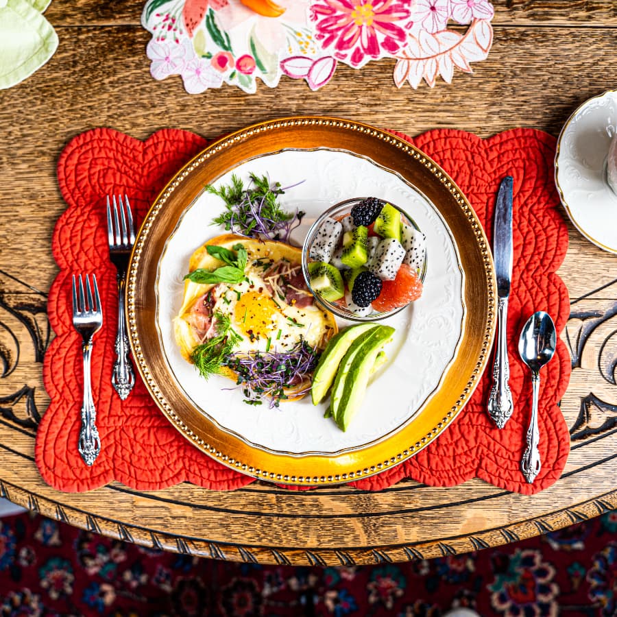 A beautifully plated breakfast featuring an egg dish with herbs, alongside a fruit cup and green apple slices, on an ornate wooden table.