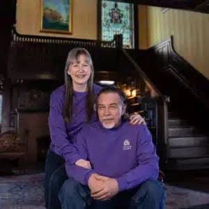 A woman and a man wearing purple sweaters pose together in a warmly lit room with a staircase in the background.