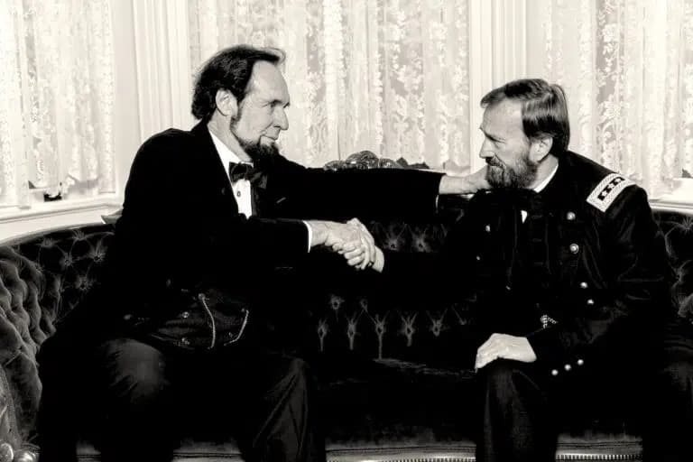 Two men in formal attire shake hands while seated on a vintage sofa.