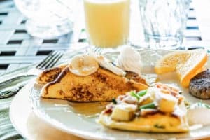 A plate of breakfast featuring a banana-filled crepe, an omelet with vegetables, and an orange slice, accompanied by a glass of juice.