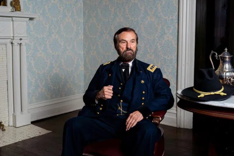 A man in a blue military uniform sits in a vintage chair in a room with patterned wallpaper.