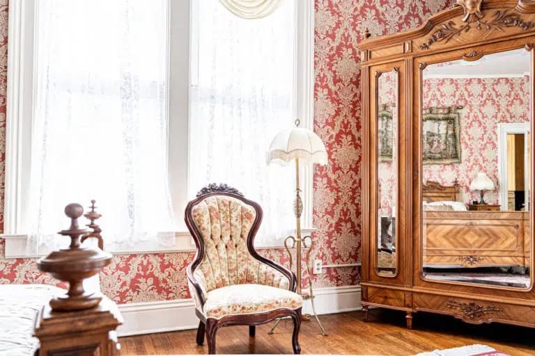 A vintage-style bedroom featuring a floral-patterned armchair, ornate wooden mirror, and elegant wallpaper.