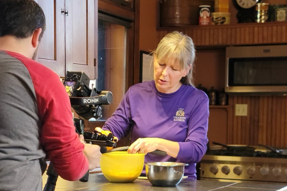 A woman in a purple shirt prepares ingredients in a kitchen while being filmed.