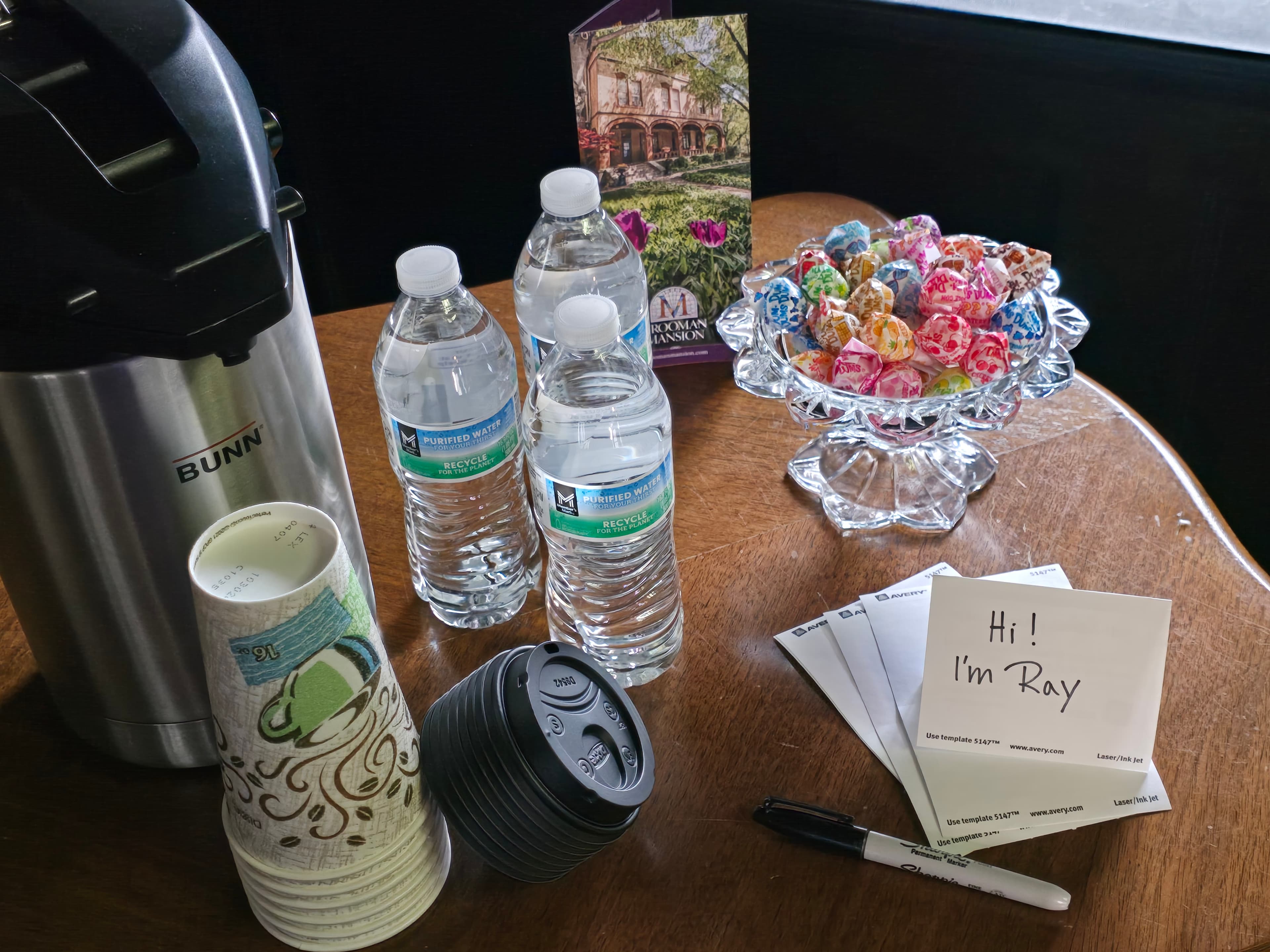 A coffee urn, water bottles, cups, a bowl of lollipops, and a note introducing "Ray" on a wooden table.
