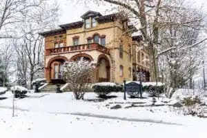 Victorian-style house covered in snow, surrounded by leafless trees and shrubs.
