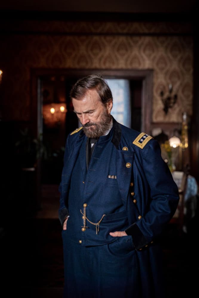 A bearded man in a blue military uniform stands contemplatively in a dimly lit, vintage interior.
