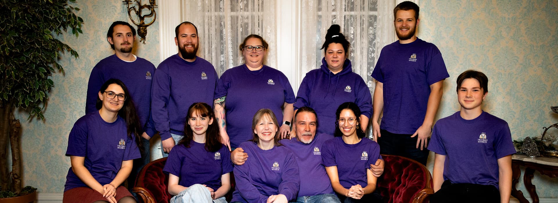 A group of eleven people wearing purple shirts poses together in a warmly decorated indoor setting.