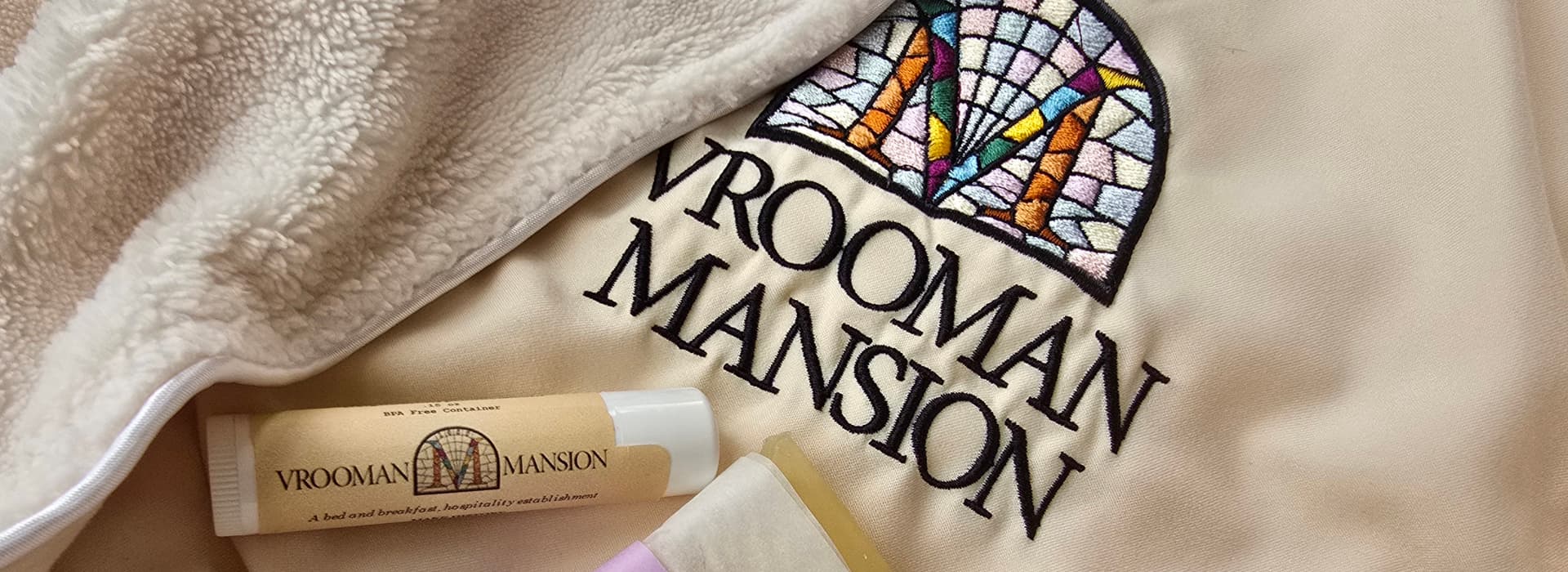 A close-up of a beige fabric featuring the embroidered logo of "Vrooman Mansion" alongside two small containers.