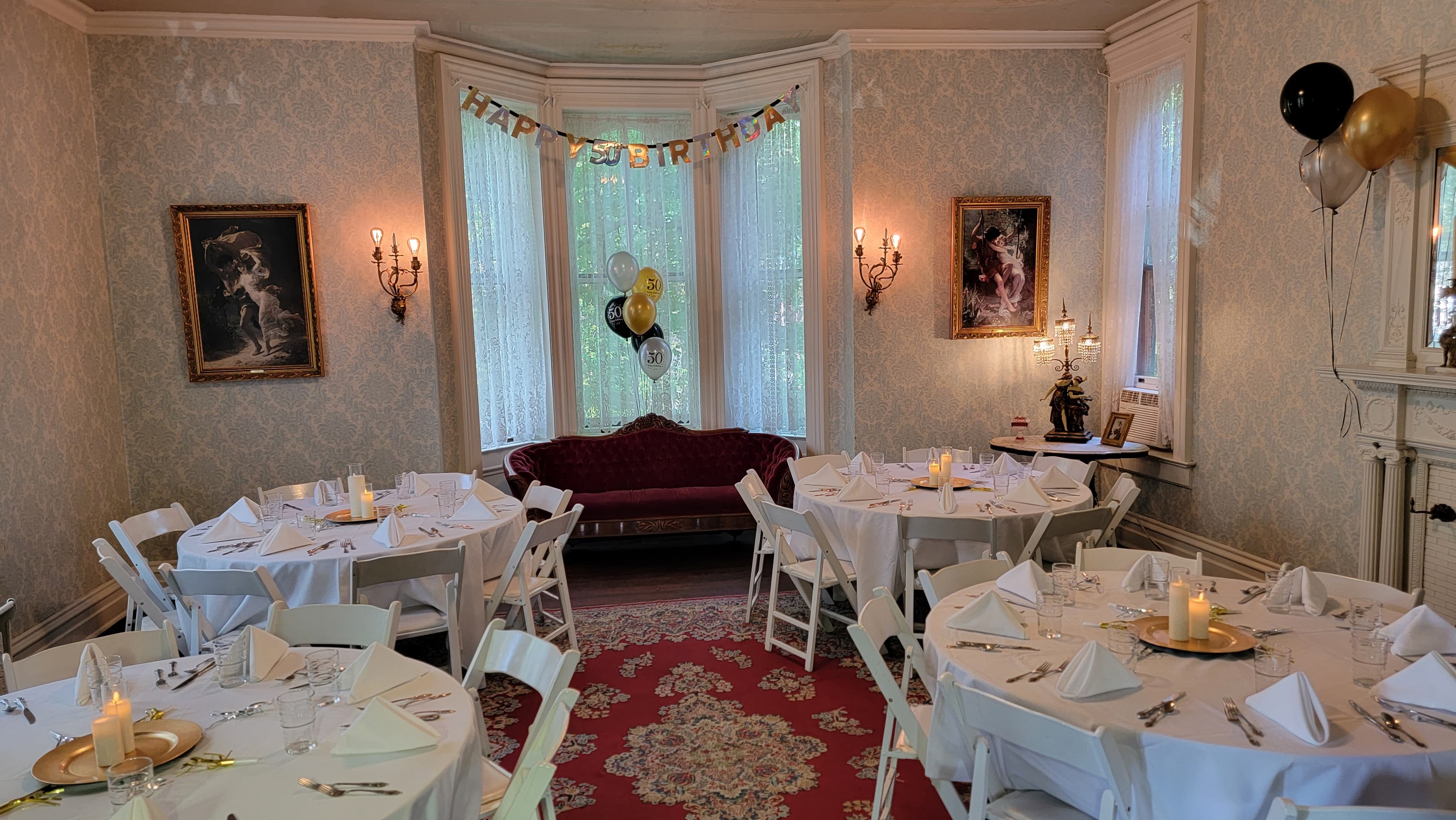 A beautifully decorated room featuring round tables with white tablecloths, a burgundy sofa, and a "Happy Birthday" banner.