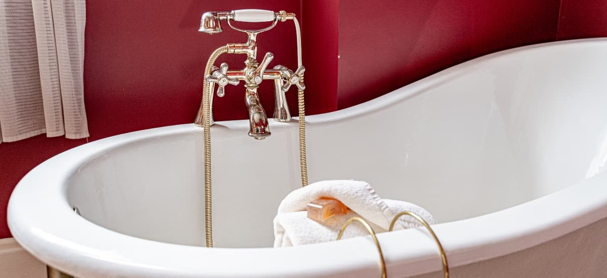 A vintage white bathtub with a gold-finished faucet and a folded towel beside it against a red wall.