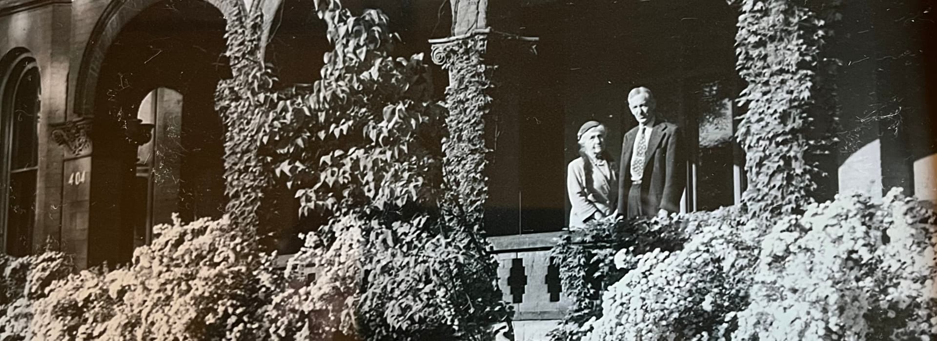 Two people stand on a balcony surrounded by lush plants.