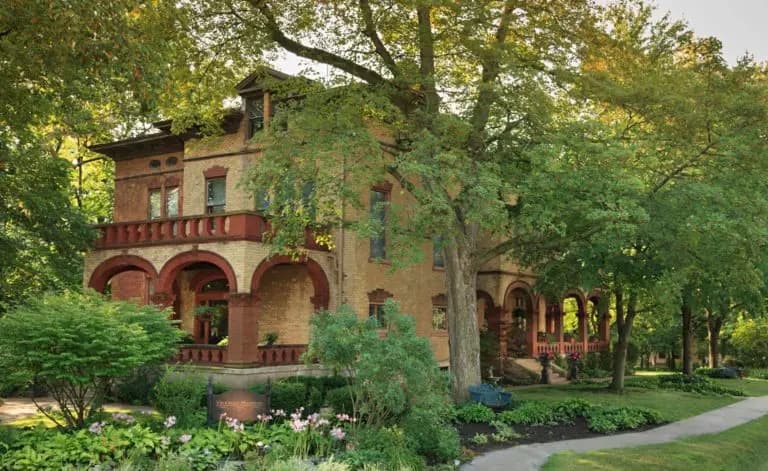 A large, historic brick house surrounded by lush greenery and blooming flowers.