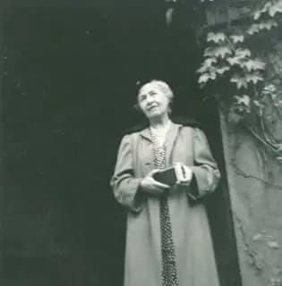 A elderly woman holding a book stands in front of a wall covered with ivy.