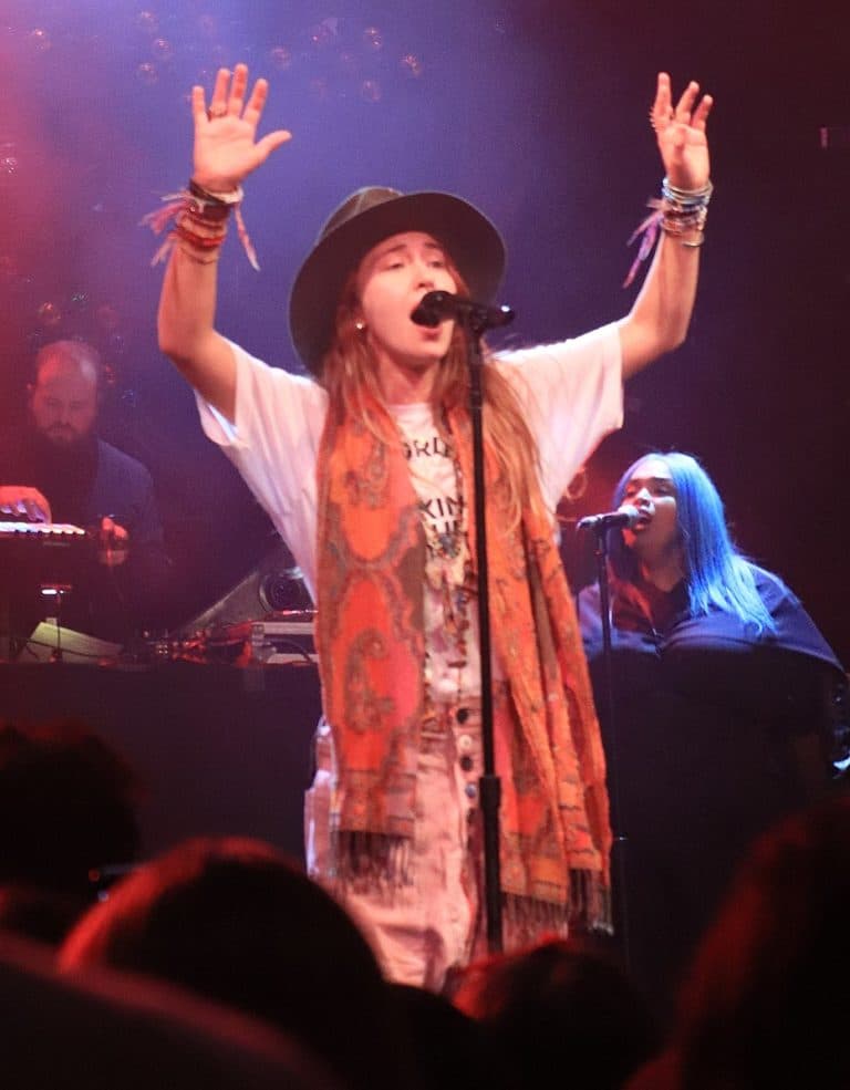 A woman wearing a t-short, a colorful orange scar, and a brown hat singing in front of a microphone with her hands raised. A woman wearing a t-short, a colorful orange scar, and a brown hat singing in front of a microphone with her hands raised.