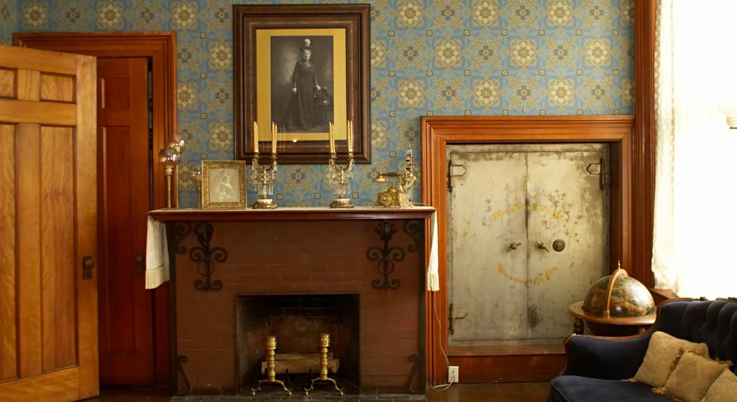 Blue papered walls, fireplace with candlesticks on the mantel and small heavy door leading to a safe room