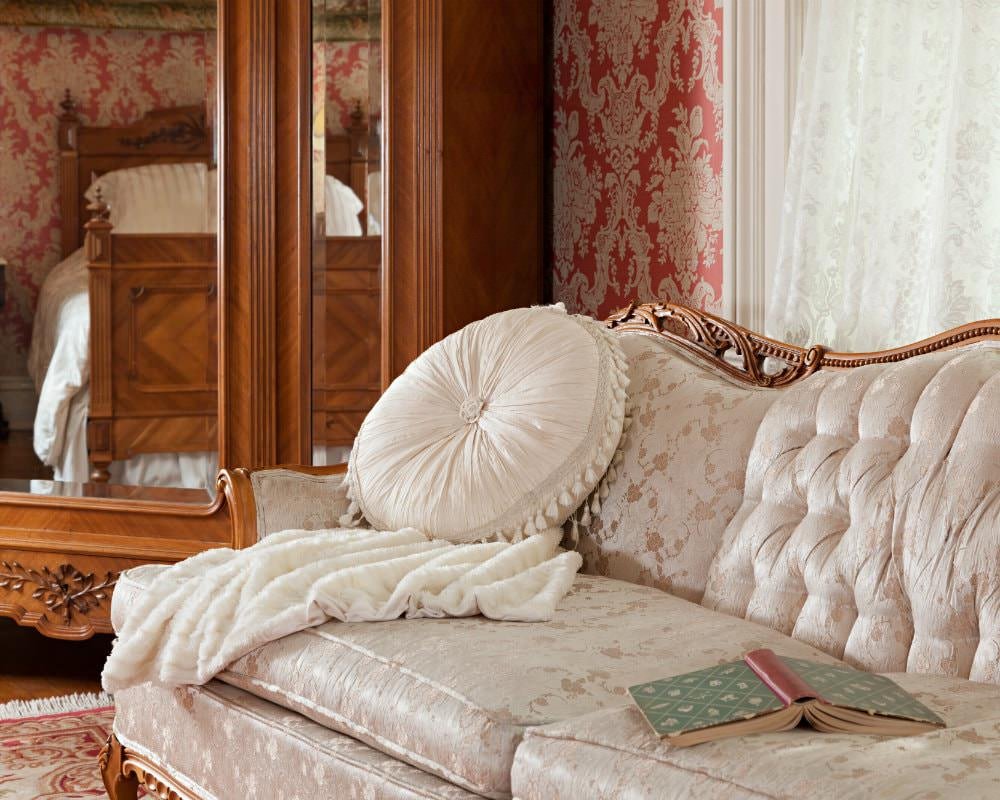 An ivory and gold sofa near a window with lace curtains and a wood mirrored cabinet