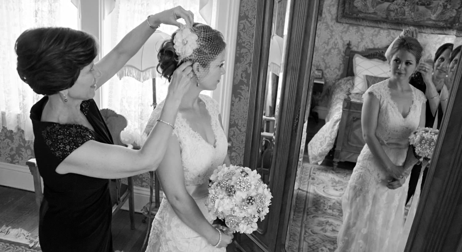 Black and white photo of bride holding bouquet in front of tall mirror while an older woman adjusts her hair
