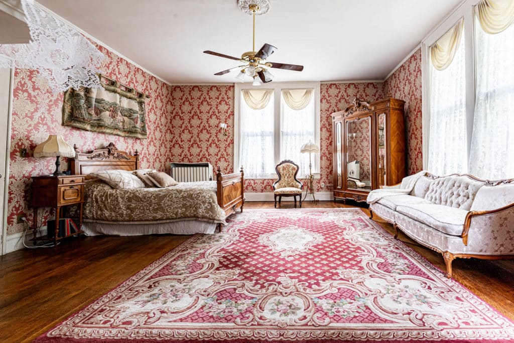 beautiful red white and pink vintage rug spans the entire room with a queen sized bed, mirrored armoire and antique sitting chair and couch in cream