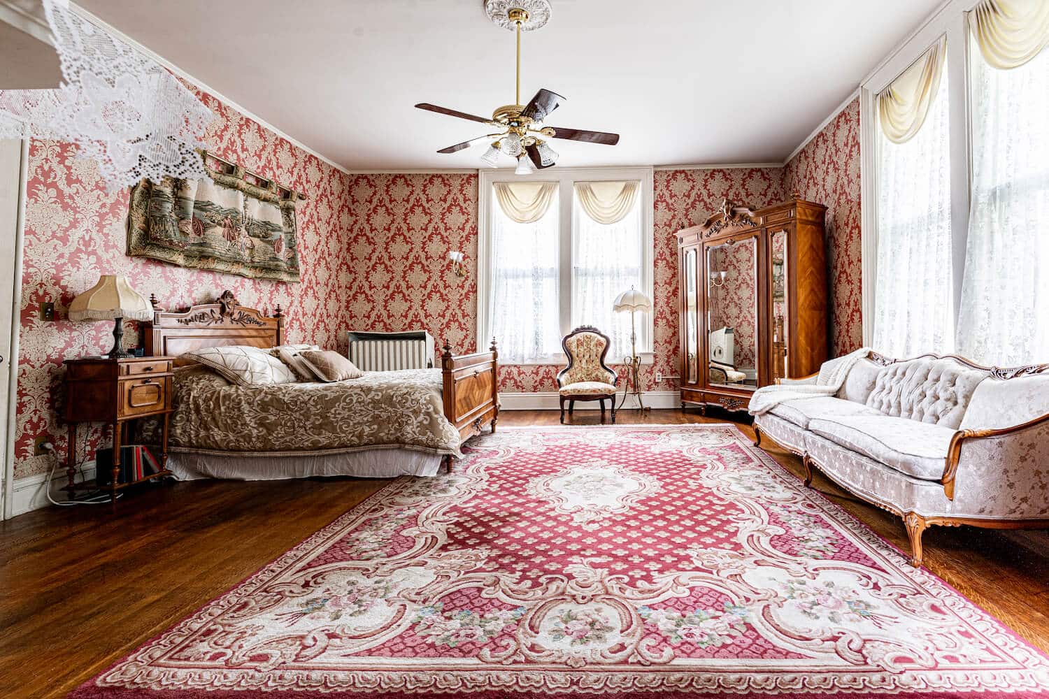 spacious room with red and white papered walls, wood floors, wood carved bed with white bedding, and large mirrored cabinet