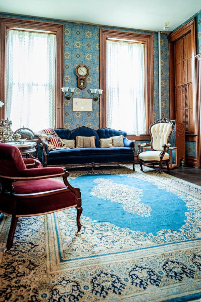 red velvet chairs and couches in the parlor with a bright red antique rug underneath, light blue wallpapered walls and silver servingware on white marble tabletops