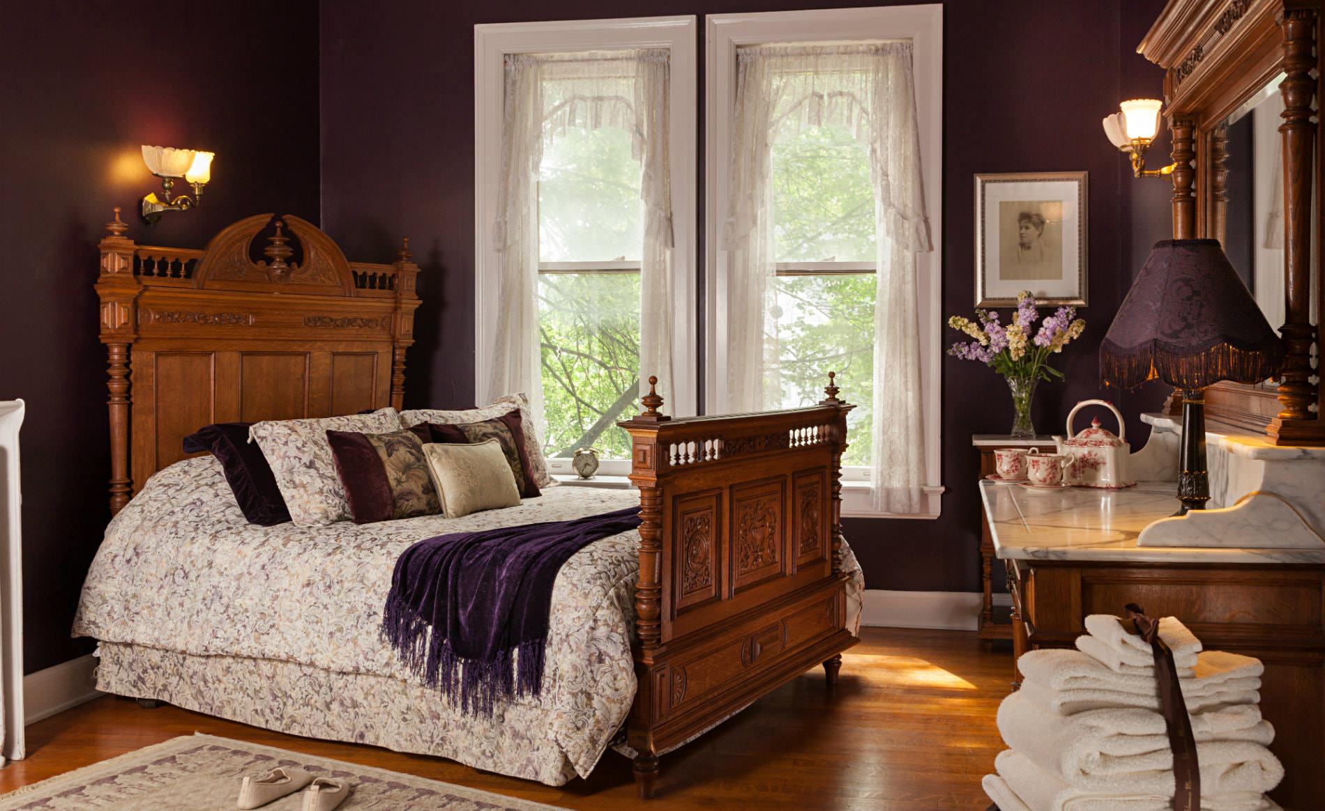 Deep purple room with white trim, white curtains, wood floors, and carved wood bed covered in floral bedding
