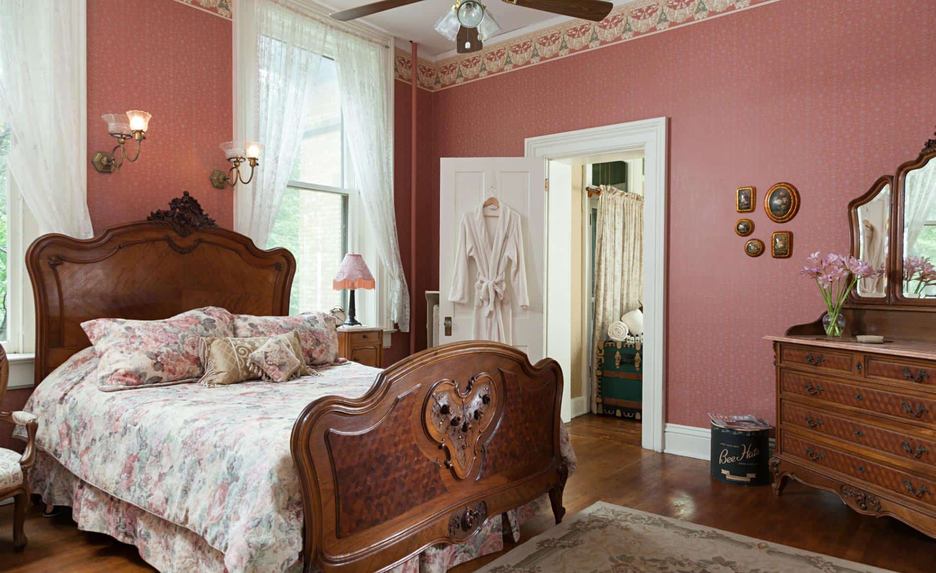 Rose papered walls, white lace curtains, white trim and doors, wood floors and elegant carved bed and dresser with mirror