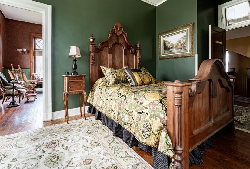 Deep green room with white trim and doors, wood floors, fireplace, and large elegant wood bed with floral bedding