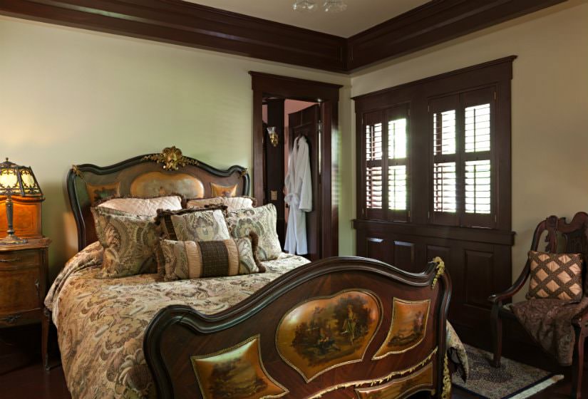 Beige room with dark stained trim and doors, wood floors, elaborate bed and a nightstand with antique lamp