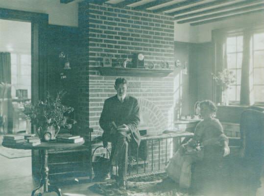 Old black and white photograph of a man and woman sitting in a sitting room near a brick fireplace