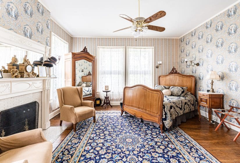Blue and beige papered walls, wood floors, white lace curtains, wood bed, mirrored cabinet and lots of natural light