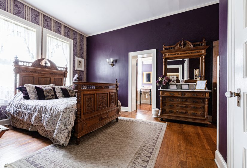 Deep purple room with white trim, white curtains, wood floors, and carved wood bed covered in floral bedding