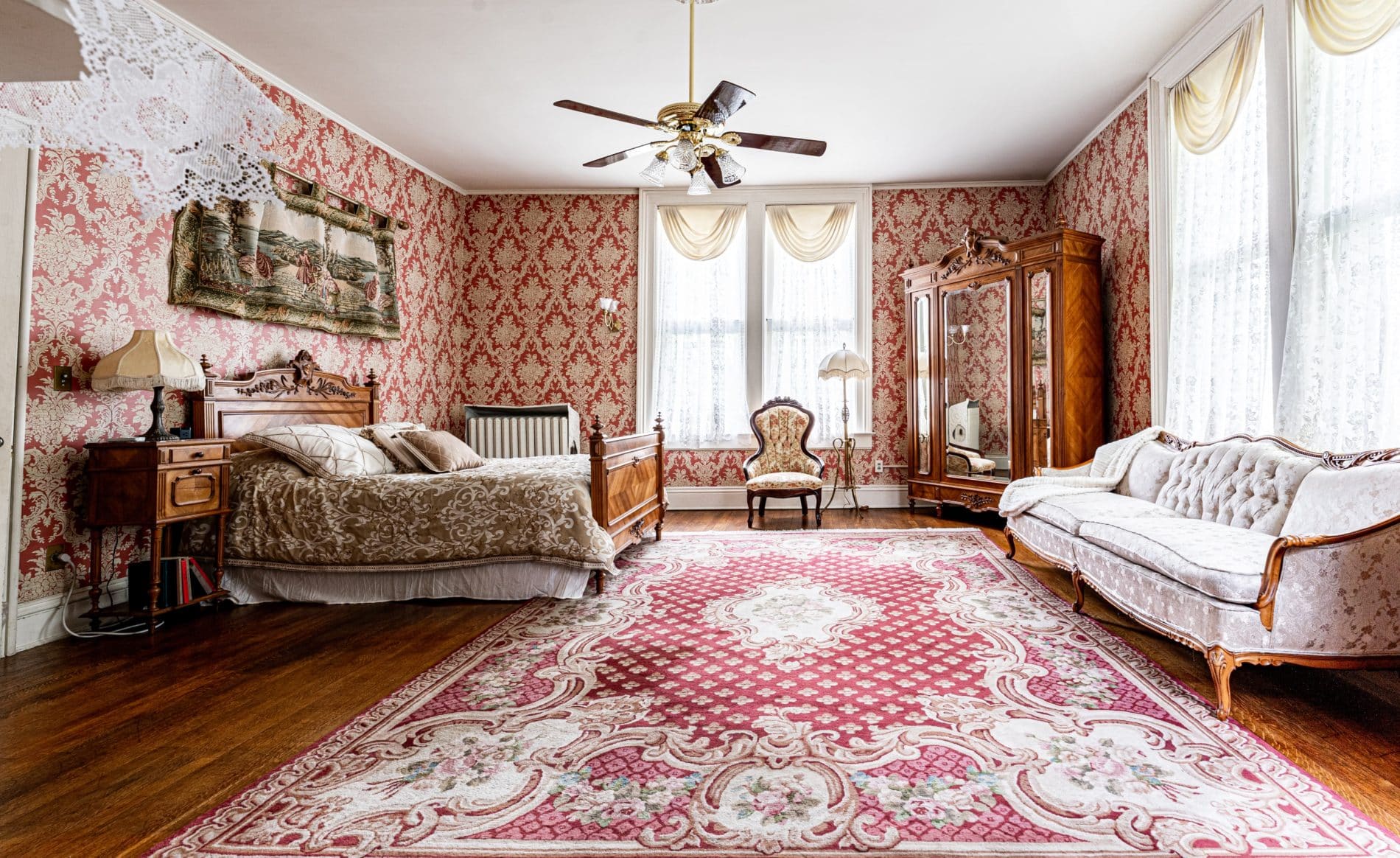 Spacious room with red and white papered walls, wood floors, wood carved bed with white bedding, and large mirrored cabinet