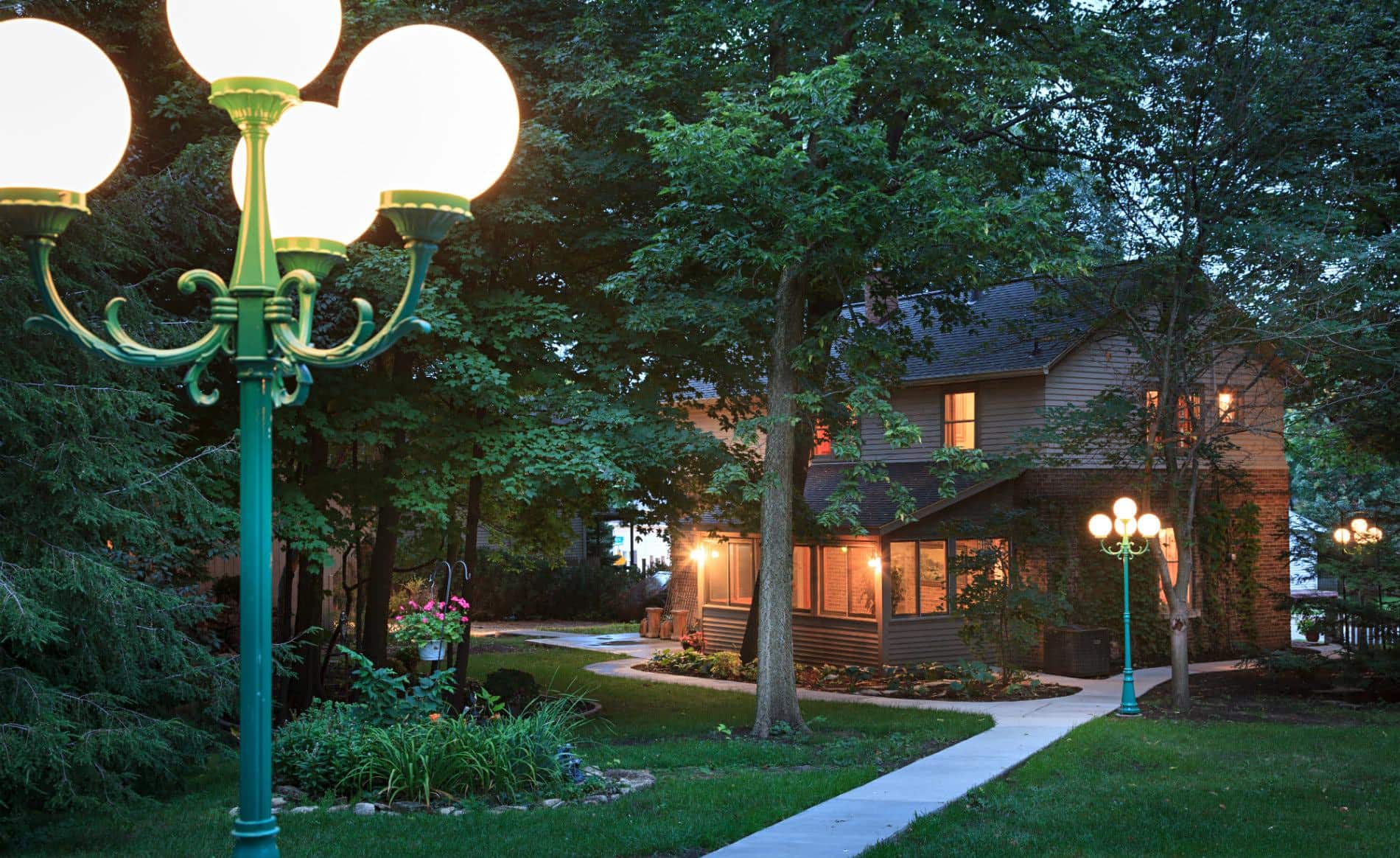 Exterior view of backyard at dusk with a sidewalk, green grass, a green light post and several trees