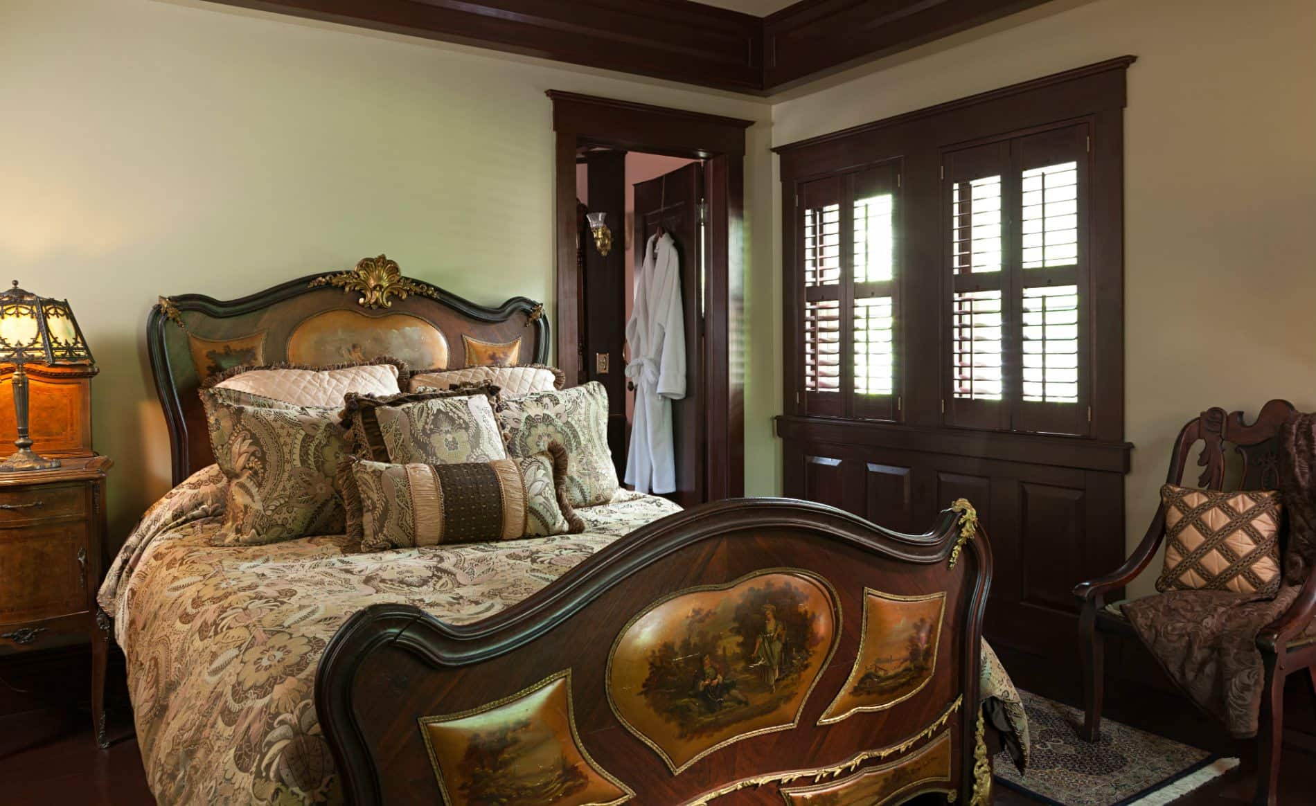 Beige room with dark stained trim and doors, wood floors, elaborate bed and a nightstand with antique lamp